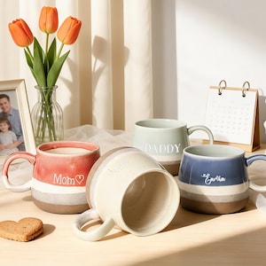 Puede incluir: Cuatro tazas de cerámica en rojo, crema, verde salvia y azul con rayas marrones y blancas. Las tazas tienen asas y las palabras "Mom", "Daddy" y "Cartha" están escritas en ellas. Un jarrón con tulipanes naranjas, una foto enmarcada y un calendario están en el fondo.