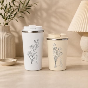 May include: A white travel tumbler with a floral line drawing and the name Leona, and a cream-coloured travel tumbler with a floral line drawing and the name Lynn. Both tumblers feature a stainless steel rim and a matching lid with a secure closure.