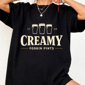 以下が含まれることがあります： クリーム色のグラフィックが施された黒いTシャツ。グラフィックは、単語「CREAMY」とフレーズ「FOOKIN PINTS」の上に3つのビールグラスが描かれています。「17」と「59」の数字がビールグラスの両側にあります。