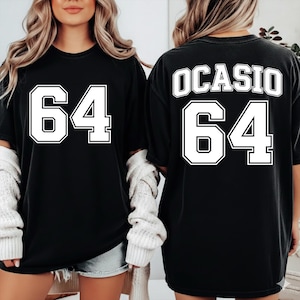 Puede incluir: Camisetas negras con el número "64" en letras blancas en la parte delantera. La parte trasera de una camiseta muestra "OCASIO" encima del número "64". Las camisetas tienen un corte holgado y de gran tamaño.