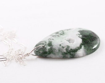 May include: A teardrop-shaped pendant necklace featuring a moss agate stone with green and white patterns. The stone is wire-wrapped and suspended from a delicate silver chain. The necklace is displayed on a white background.