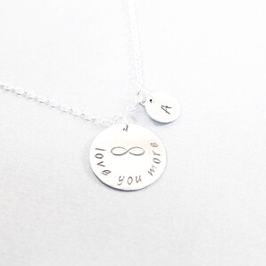 May include: A silver necklace with two pendants. The larger pendant is a circle with the words "love you more" and an infinity symbol. The smaller pendant has the letter "A". The necklace is on a white background.