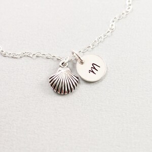May include: A silver necklace featuring a seashell charm and a small circular pendant with the letter "M" in black script. The chain is delicate, and the seashell charm has detailed ridges.