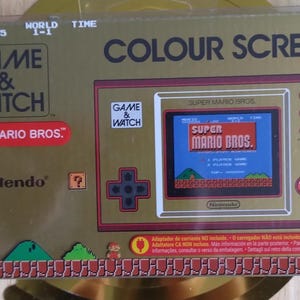 Könnte beinhalten: Eine goldfarbene Game & Watch Super Mario Bros. Handheld-Konsole. Die Box zeigt den Spieletitel, einen Farbbildschirm und das Nintendo-Logo. Der Bildschirm des Spiels zeigt ein klassisches Super Mario Bros. Level.