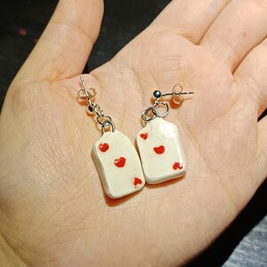 Può includere: Un paio di orecchini fatti a mano con un design rettangolare bianco e dettagli a forma di cuore rosso. Gli orecchini sono sospesi a ganci e perni color argento. Gli orecchini sono esposti su uno sfondo chiaro.