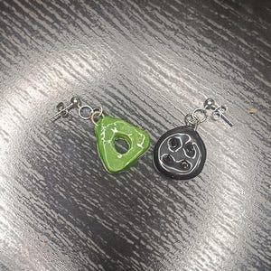 Può includere: Un paio di orecchini fatti a mano. Un orecchino è un triangolo verde con un foro al centro. L'altro orecchino è un cerchio nero con un disegno bianco. Entrambi gli orecchini hanno perni e anelli color argento.