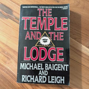 Peut inclure: Un livre intitulé "The Temple and the Lodge" de Michael Baigent et Richard Leigh. La couverture est noire avec du texte rouge et blanc. Un triangle avec un œil est au centre. Le livre contient une citation de Booklist.