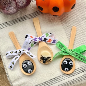 May include: Three wooden spoons decorated for Halloween. Each spoon has a different design: one with googly eyes, one with a spider, and one with googly eyes. The spoons are tied with decorative ribbons. A glittery purple pumpkin and an orange pumpkin are in the background.