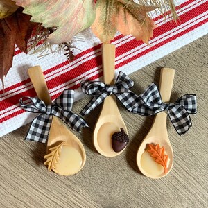 May include: Three wooden spoons with decorative resin and gingham bows. Each spoon features a different fall-themed design: an acorn, an oak leaf, and a maple leaf. The spoons are arranged on a wooden surface with a red and white striped cloth.