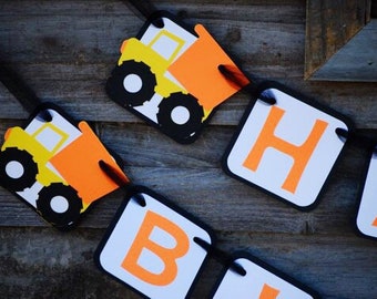 May include: A banner with construction truck and letter cutouts. The trucks are yellow and orange with black wheels. The letters are orange on white backgrounds. The banner is strung on black string and is set against a wooden background.