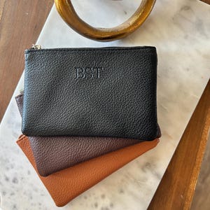 May include: A stack of three small leather coin purses in black, brown, and tan. The top black purse has the embossed initials "BST" and a silver zipper. The purses are stacked on a marble surface.