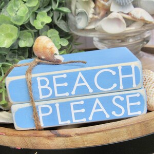 May include: Two blue wooden blocks stacked on top of each other. The top block says "BEACH" in white letters and the bottom block says "PLEASE" in white letters. The blocks are tied together with twine and a seashell is on top of the twine.
