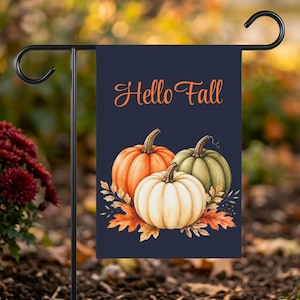 以下が含まれることがあります： 濃いネイビーブルーの背景にオレンジ色の「Hello Fall」という文字が上部に書かれた長方形のガーデンフラッグ。文字の下には、オレンジ、白、くすんだ緑色の3つのカボチャが描かれ、その周りをオレンジと茶色の秋の葉が囲んでいます。