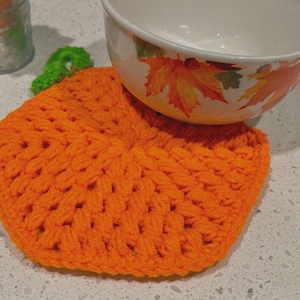 May include: An orange, crocheted pumpkin-shaped trivet with a green stem. A white bowl with fall leaf designs rests on top. The trivet is handmade and likely used for kitchen or dining decor, adding a seasonal touch.