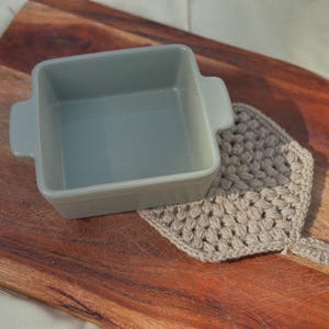 May include: A square, light gray ceramic baking dish with handles sits on a wooden cutting board next to a beige crocheted potholder. The dish is empty and ready for use. The potholder is handmade.