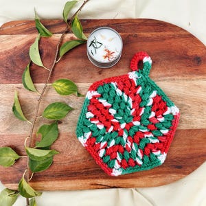 May include: A handmade, crocheted hot pad in a vibrant red, green, and white color scheme. The hexagonal pad features a loop for hanging and is placed on a wooden cutting board. A small candle in a metal tin is also present.