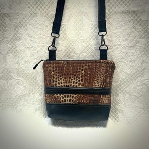 May include: A crossbody bag with a brown and black reptile print pattern. The bag has a black leather bottom and a black adjustable strap. The bag has a zipper closure and black hardware.