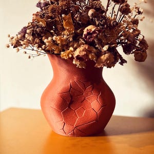 May include: A terracotta-colored vase with a geometric pattern holds a bouquet of dried flowers. The flowers are a mix of brown, purple, and cream hues, creating a rustic and natural aesthetic. The vase has a textured surface.