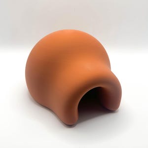May include: A terracotta-colored, rounded object with a curved opening. The smooth, matte surface has a uniform color. The shape is reminiscent of a stylized head or a unique decorative piece.