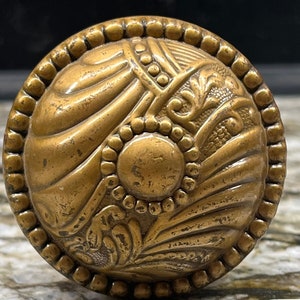 May include: A vintage brass door knob with an ornate design. The knob is approximately 1 1/2 inches in diameter and has a raised, circular center. The knob is surrounded by a beaded border and features a floral pattern. The knob is marked "MADE IN U.S.A." and "PAT. NO. 2,562,335".