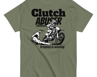 May include: An olive green t-shirt featuring a black graphic of a person riding a dirt bike, with the text "Clutch ABUSER", "WHEELIEWORKS", and "Dripping is winning" printed in black on the front center of the garment.