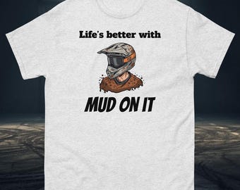 May include: Light grey t-shirt with the text "Life's better with MUD ON IT." The shirt features a graphic of a motocross helmet and a person's head. The helmet is brown and orange.