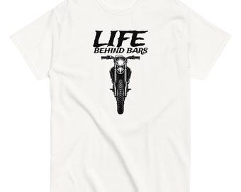 May include: A white short-sleeve t-shirt featuring a black graphic of a motorcycle viewed from the front. Above the motorcycle, the text reads "LIFE BEHIND BARS" in a bold, stylized black font. The design is centered on the chest of the shirt.