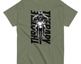May include: An olive green t-shirt featuring a black and white graphic of a motocross rider on a dirt bike. The text "THROTTLE THERAPY" is printed vertically in bold black capital letters on both sides of the rider illustration.
