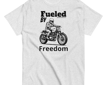 May include: A light gray t-shirt featuring a black graphic of a person wearing a helmet riding a motorcycle. The text "Fueled BY" appears above the rider, "WheelieWorks" is in small print, and the word "Freedom" is printed below the motorcycle.