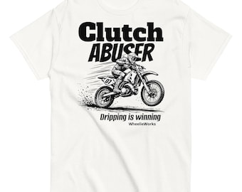 May include: A white t-shirt featuring a black graphic of a motocross rider on a dirt bike with the number 07. The text reads Clutch ABUSER, Dripping is winning, and WheelieWorks. The design depicts motion lines and dirt spray behind the motorcycle.