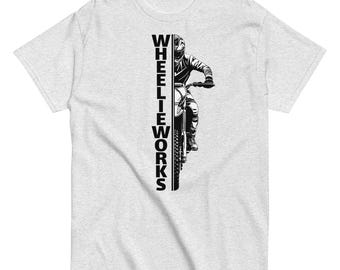 May include: A light gray short-sleeve t-shirt featuring a black graphic print. The design displays a vertical line of text reading WHEELIEWORKS next to a black and white illustration of a person riding a dirt bike in a wheelie position.
