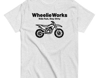 May include: A light grey short-sleeve t-shirt featuring a black graphic of a dirt bike. Above the bike, the text reads "Wheelie Works" and below the bike, the text reads "Ride Fast, Stay Dirty" in a bold, black, sans-serif font.