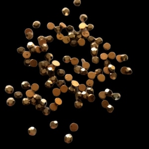 May include: A scattering of small, metallic gold-colored beads against a black background. The beads have a faceted, gem-like appearance, reflecting light. These decorative beads are likely used for embellishing crafts or jewelry.