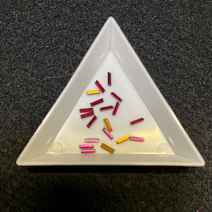 May include: A white triangular tray filled with small, colorful, rectangular rhinestones. The rhinestones are in shades of pink, red, and gold.