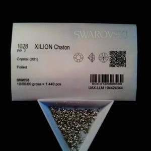 May include: A close-up of a small, white, triangular tray filled with silver-colored Xilion Chaton crystals. The tray is labeled with the product number 1028, the name "XILION Chaton", and the size "PP 7". The label also includes the text "Crystal (001)", "Foiled", and "SWAROVSKI".