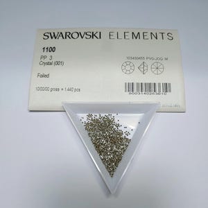 Puede incluir: Una bandeja triangular blanca llena de pequeños cristales brillantes, junto a una tarjeta blanca con el texto "SWAROVSKI ELEMENTS". La tarjeta también muestra información del producto, incluyendo "1100 PP 3 Crystal (001) Foiled".