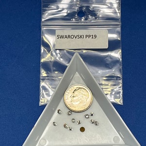 Puede incluir: Una bandeja triangular blanca con un pequeño montón de piedras de cristal transparentes y una moneda de 10 céntimos de euro. La bandeja está etiquetada con "SWAROVSKI PP19" en una etiqueta blanca.