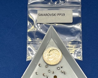 May include: A white triangular tray with a small pile of clear rhinestones and a US dime coin. The tray is labeled "SWAROVSKI PP19" on a white label.