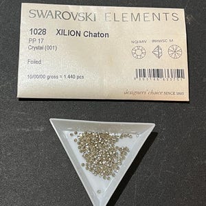 May include: A white triangular tray filled with many small, sparkling crystal rhinestones. Above the tray is a card labelled "SWAROVSKI ELEMENTS", with text including "XILION Chaton" and "Crystal (001)". The card also has a barcode and the text "designers' choice SINCE 1893".