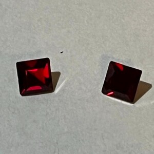 Puede incluir: Dos gemas cuadradas de color rojo intenso. Las gemas están sobre un fondo claro, con sombras que sugieren una fuente de luz. Las facetas de las piedras capturan la luz, creando un brillo brillante. Para joyería.