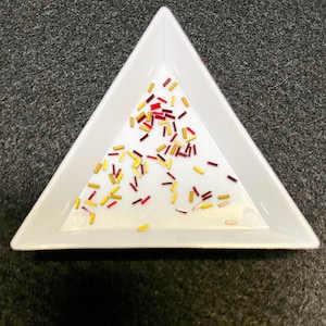 May include: A white triangular tray filled with small, colorful glass beads. The beads are red, gold, and white.