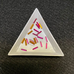 以下が含まれることがあります： ピンクとゴールドの四角いラインストーンが詰まった白い三角形のトレイ