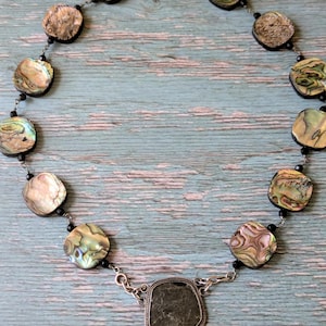 May include: Abalone shell necklace with a large, square, dark green stone pendant. The necklace features iridescent, round abalone shell beads and small black beads strung on a silver chain. The pendant is framed in a silver setting.