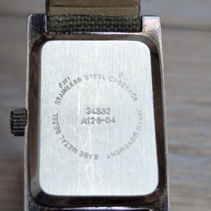 Puede incluir: Primer plano de la parte trasera de un reloj rectangular con un bisel de metal plateado y una correa de lona verde. La esfera del reloj es plateada con el texto "STAINLESS STEEL CASEBACK", "JAPAN MOVEMENT" y los n&uacute;meros "24832 A126-04".
