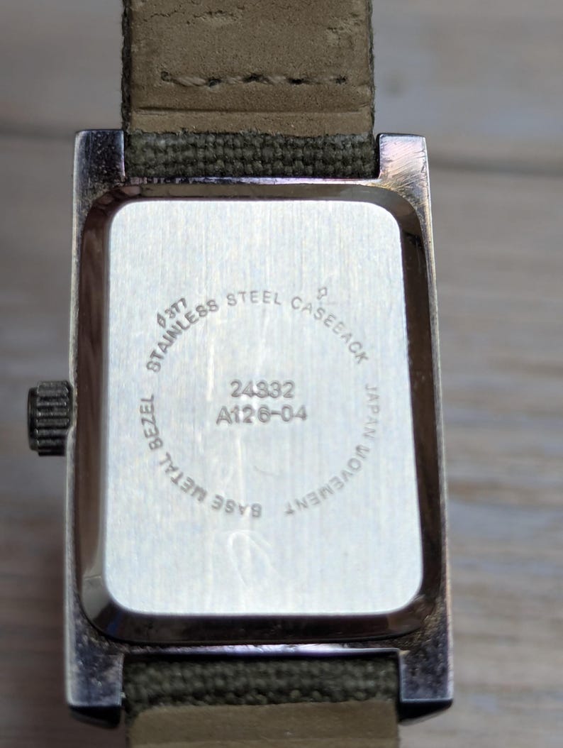 Puede incluir: Primer plano de la parte trasera de un reloj rectangular con un bisel de metal plateado y una correa de lona verde. La esfera del reloj es plateada con el texto "STAINLESS STEEL CASEBACK", "JAPAN MOVEMENT" y los n&uacute;meros "24832 A126-04".