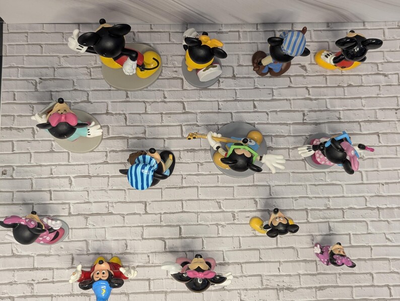 May include: A collection of Mickey Mouse figurines in various poses, each mounted on a circular base. The figurines feature Mickey in different outfits, with vibrant colors like red, yellow, and blue. The figures are displayed against a white brick wall.