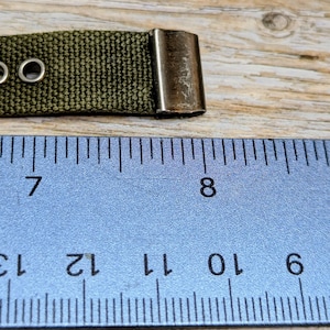 Puede incluir: Primer plano de una correa de lona verde con dos ojales met&aacute;licos y un cierre met&aacute;lico. Una regla azul con marcas y n&uacute;meros negros est&aacute; en primer plano. La correa mide aproximadamente 2,5 cm de ancho.