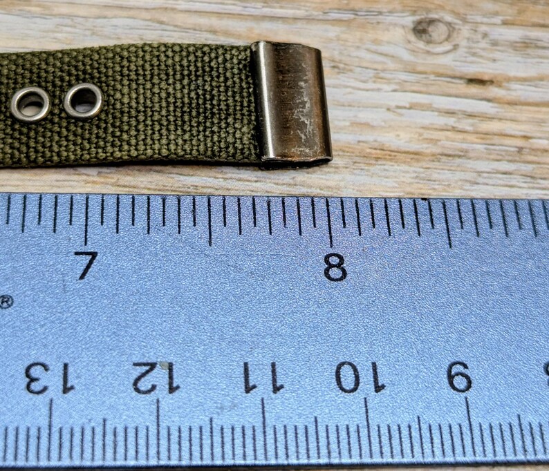 Puede incluir: Primer plano de una correa de lona verde con dos ojales met&aacute;licos y un cierre met&aacute;lico. Una regla azul con marcas y n&uacute;meros negros est&aacute; en primer plano. La correa mide aproximadamente 2,5 cm de ancho.