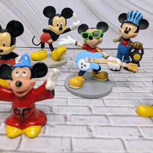 May include: A collection of Mickey Mouse figurines in various poses and outfits. The figurines include Mickey as a conductor, a guitarist, and a magician, all with yellow shoes and black ears. The figures are on a white brick surface.
