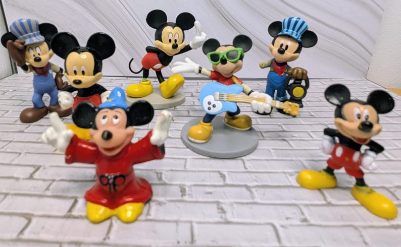 May include: A collection of Mickey Mouse figurines in various poses and outfits. The figurines include Mickey as a conductor, a guitarist, and a magician, all with yellow shoes and black ears. The figures are on a white brick surface.